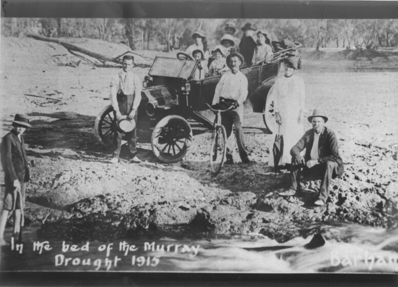 Black and white photograph of a car load of people parked in a dry river bed. Some men and women stand and sit nearby the car. Large gum trees are visible in the background.