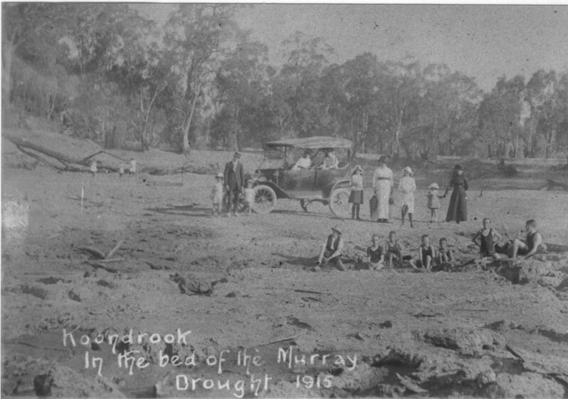 Black and white photograph of a group of people sitting and standing in the dried river bed. A group of children sit on the ground in a row in bathing suits, while behind them, a dressed up family stand in front of their car, holding hands and looking towards the camera. Large gum trees stand on what would have been the riverbank.