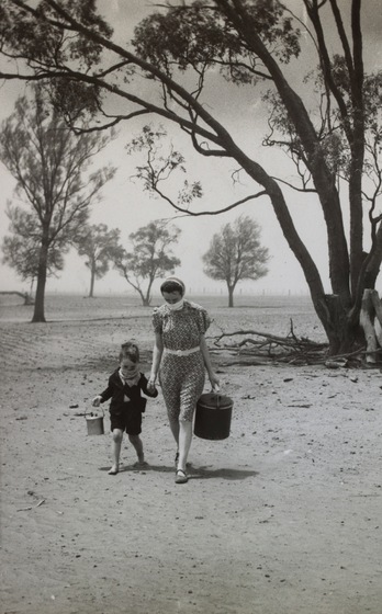 A mother and child walk through a dry barren landscape, each holding a pot in their hand and a scarf wrapped around their mouths to stop the dust. Dead trees stand in the distance.