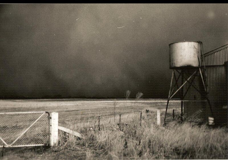 Landscape of a very dark sky, made up of dust, stretching across the horizon. In the foreground in a wire fence and gate and a water tank of a stand next to a corrugated shed.