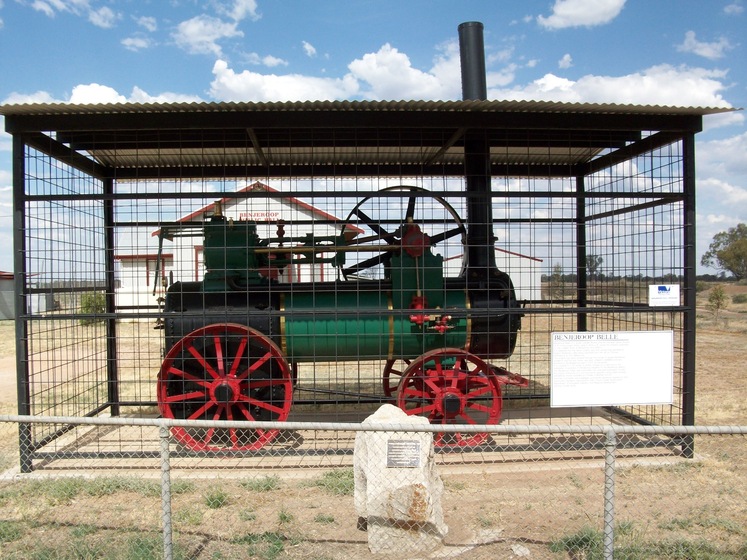 An old green, black and red steam engine enclosed in a wire cage in an open, dry park. A memorial stone sits at the base of the train and an information board is attached to the wire fence in front of the cage.