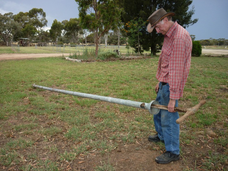 An older man in a checked shirt and jeans holds a long metal instrument that is resting on the ground. The man stares down at the instrument, he is holding it by a timber handle.