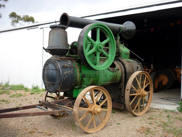 A black and green steam engine, with yellow wheels, is propped up outside a large metal shed.