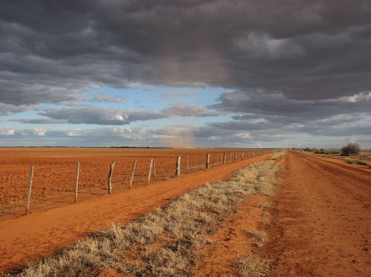 Landscape photograph of a red sandy paddock, a wire fence running down the centre of the image. The horizon is dotted with black clouds and gaps of blue and white sky.