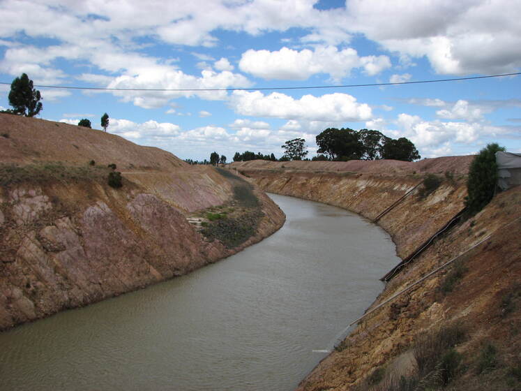 A flat bed of water runs through a channel of land, red sandy cliffs on either side and large green trees in the distance.