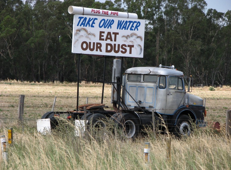 An old truck and trailer is parked next to a fence line, a large sign perched off the back of the truck with the wording 'Take Our Water and Eat Our Dust'.