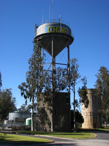 A tall metal water tower with the wording 'Cohuna' printed in large letters on the top. The tower is close to other structures, including a smaller cement tank and a tin tank.