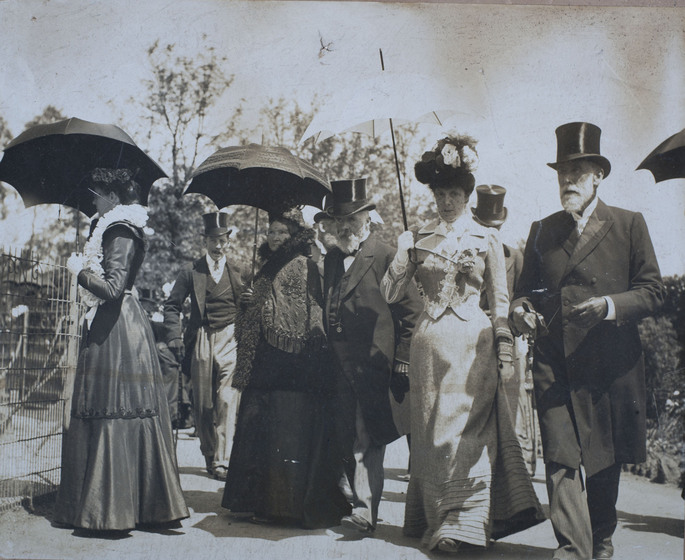 A group of well dressed men and women in suits, top hats and full length gowns and hats, stroll through the zoo grounds. Some are holding black and white parasols over their heads.