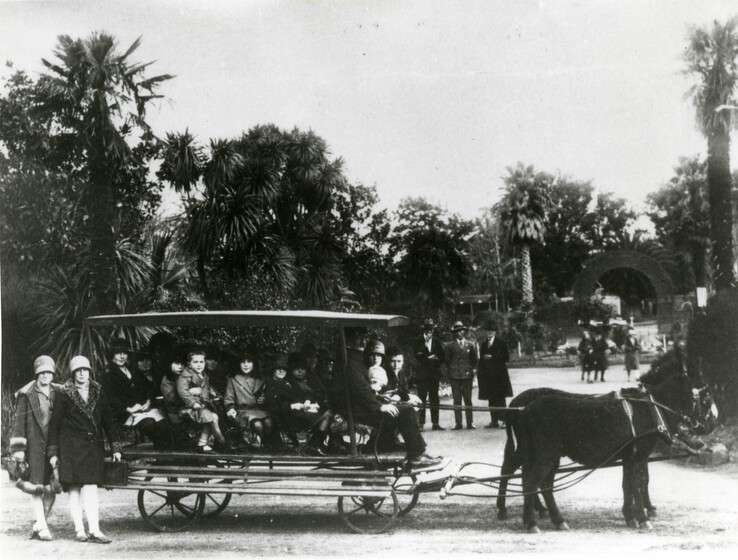 Black and white photograph of a group of children and adults sitting in a covered carriage being pulled by two donkeys. Around them are groups of people walking around the zoo grounds.