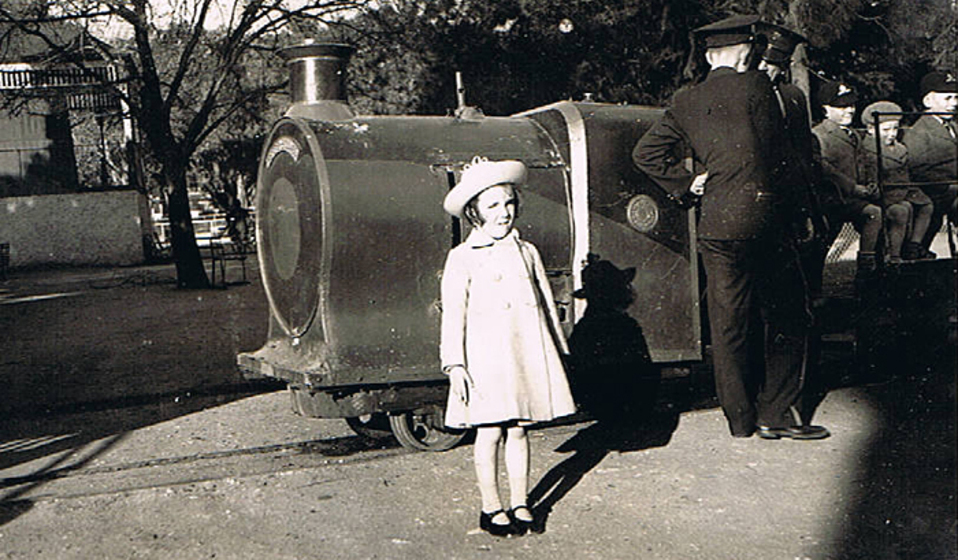 A young child in a dress and hat stands in front of a small steam engine. Two men in uniform stand behind the girl, leaning partially on the train engine.
