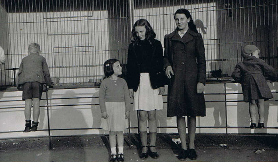 A young girl stands next to two older teenage girls in front of what appears to be a bird cage. Two small kids are behind them, leaning to look into the bird cage.
