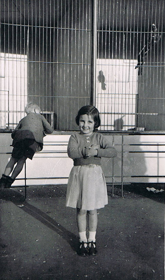 A small child in a dress stands in front of a bird enclosure, a large smile on her face. A small boy stands behind her leaning over the railings to look into the bird cage.