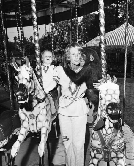 A woman stands on a carousel in between two horses. On one side of her she is partially holding a young chimp that is standing on a horse but has its arms wrapped around the ladys neck. On the other side is a small child, sitting on the horse and holding onto the ride and the lady's arm. 