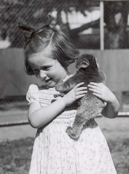 A small girl in a dress and bows in her hair holds a koala joey against her shoulder, hugging it to her.