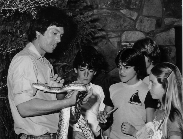 A man stands in front of a group of four children, about the age of 10, holding a long python in his arms.