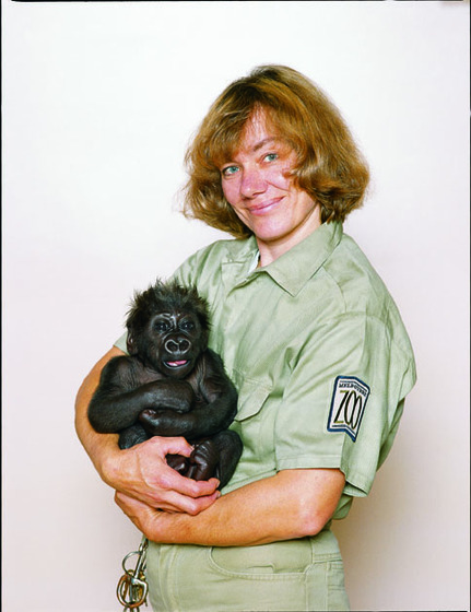 A woman in a khaki uniform holds a small baby gorilla in her arms - both are staring straight at the camera.