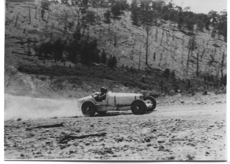 Black and white photograph of a small sports car driving across a rocky landscape with sheer rocks cliffs positioned behind them. The car is throwing up dust as it drives past.