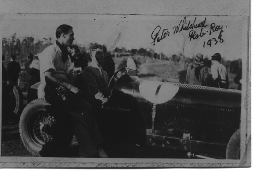 Black and white photograph of two men sitting in a sports car - one in the driving seat, and the other sitting on the tire guard. Groups of other men stand in the background behind the car.