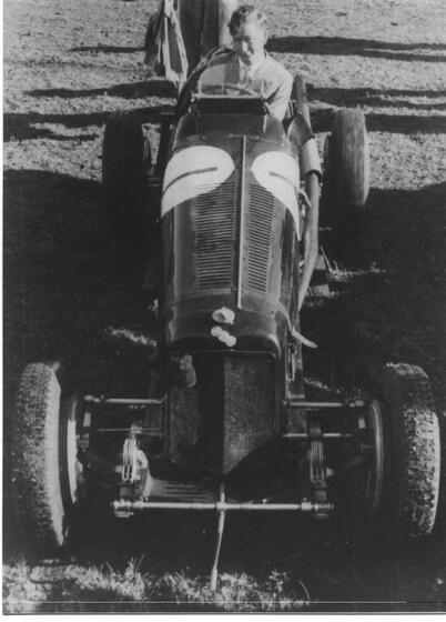 A man sits in a sports car, the long nose of the front of the car facing towards the camera. On the right hand side of the radiator sits a small clip on koala.