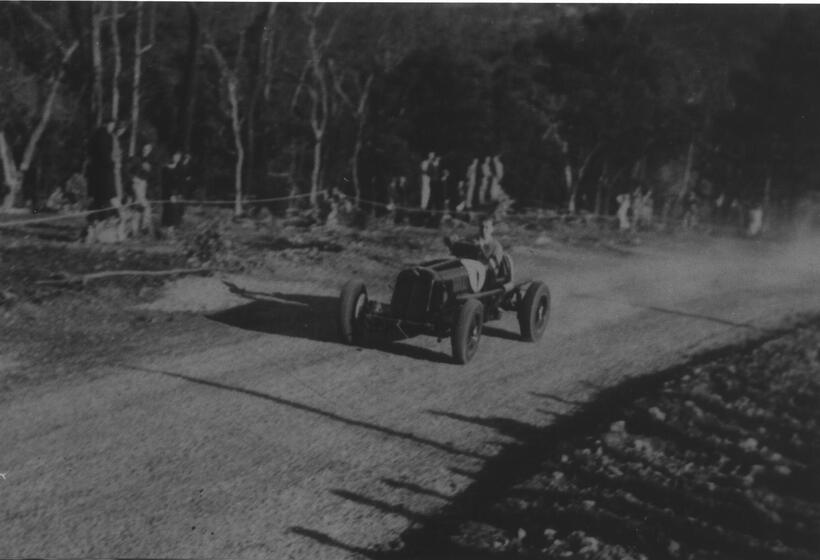Black and white photograph of a man sitting in a motorcar, driving towards the camera on a dirt track. Dust flies behind the car and spectators stand on the side of the track watching, behind a rope fence.