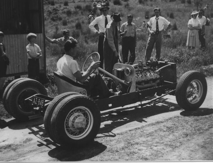 Black and white photograph of a man sitting in a stationary motorcar. The bonnet has been removed and the engine is exposed. Men, women and children stand behind the car, on the grassy verge, and observe the man in the car.