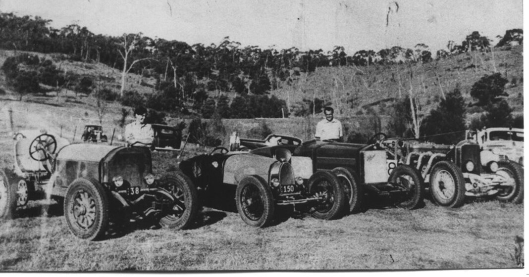 Black and white photograph of five motorcars lined up in row, in and open field. Two men stand near the cars, each leaning on one. In the distance a hills with trees covering them.