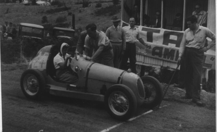 Black and white photograph of a woman sitting in a motorcar, it parked behind a white starting line. A man leans over her, both hands leaning on the bonnet, whilst another man stands on the starting line with a pole in his hand, resting on the white line. Other men stand around and observe the happenings of the start of the race.