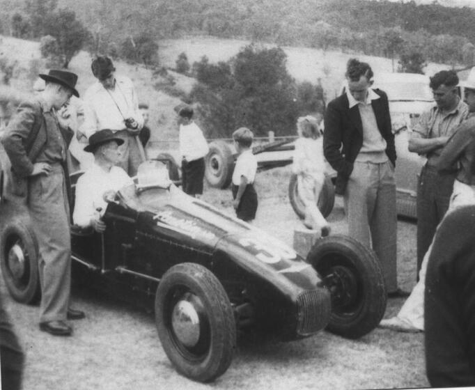 Black and white photograph of an older man sitting in a motorcar, whilst other men stand close by observing the car in some detail. Behind them children walk around looking at other things happening in the field.