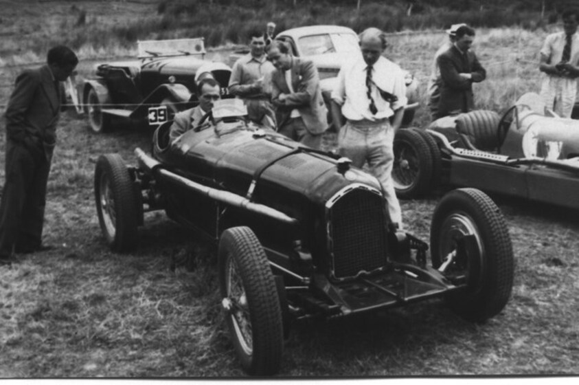 Black and white photograph of an older man sitting in a motorcar, whilst other men stand close by observing the car in some detail. Behind them are more motorcars and other men looking intently at the vehicles.