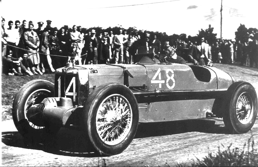 Black and white photograph of a man sitting in a race car, sunglasses on and staring intently ahead. A large group of spectators stand behind a rope fence, watching the cars go past.