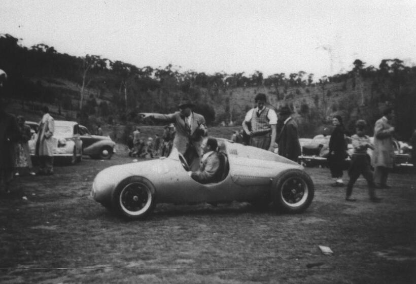 Black and white photograph of a man sitting in a sports car, parked in the middle of an open field. Other men stand close by watching, including one who is close to the car and pointing into the distance. Other people walk around the field looking at other parked cars.
