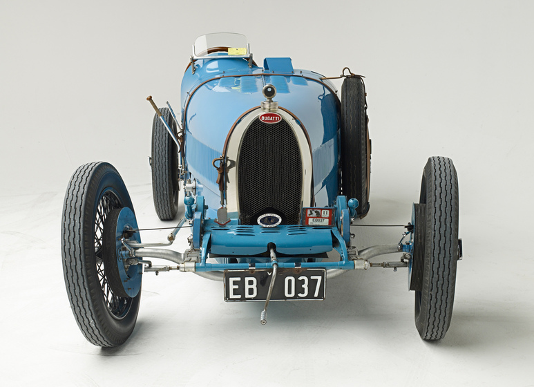 A light blue motor car with an open top, positioned with the bonnet facing the front. The front wheels stick out somewhat further from the back wheels.
