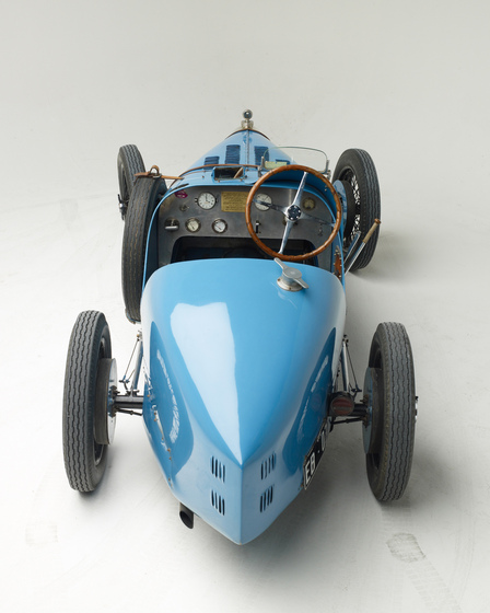 A light blue motor car with an open top, positioned with the boot facing the front. From this angle you can see the steering wheel, the dashboard and a spare tire attached to the side of the car.