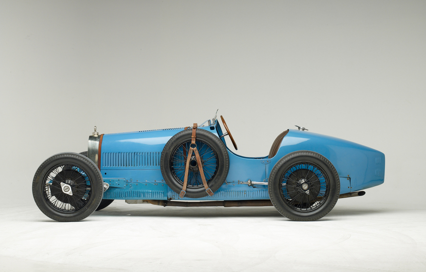 A light blue motor car with an open top, positioned with the side of the car facing the front. From this angle you can see a spare tire attached to the side, and how the front wheel is slightly larger the back wheel.