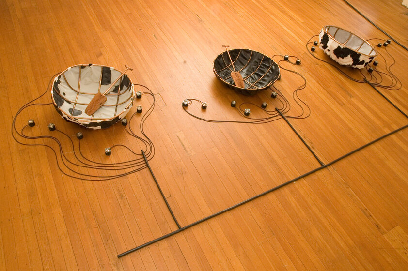 Three bowl shape structures sit on the floor - they are covered with cow hide and have black strings with some kind of weight attached, trailing off from the sides. In each rests a small wooden paddle.