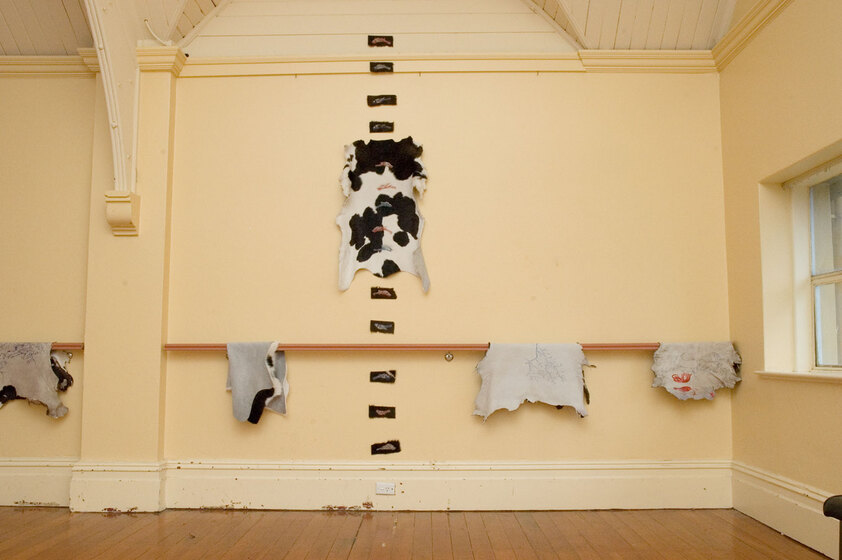 A selection of cow hides hang on a bar that is wrapped around a large room. On one part of the wall is a series of small artworks, a selection of birds etched on to cow hide, stretching up in a line towards the roof. About halfway up the row of artworks is a larger black and white cow hide positioned onto the wall.