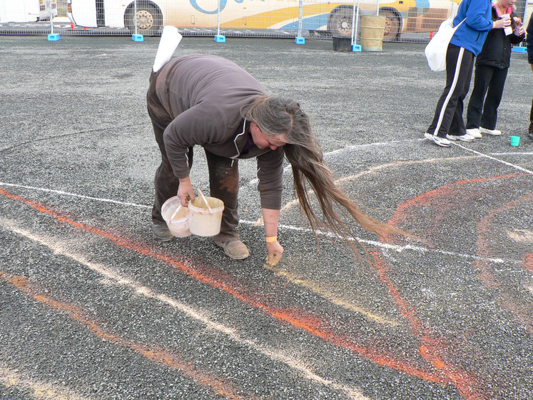 A woman leans over a bitumen surface and sprinkles sand into a row. Around her are lines of orange and white sands.
