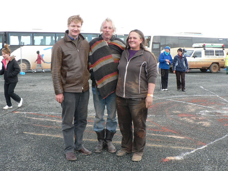 Three people stand together, there arms around each other, smiling at the camera. Around them on the bitumen ground are sand patterns and people observing the artwork.