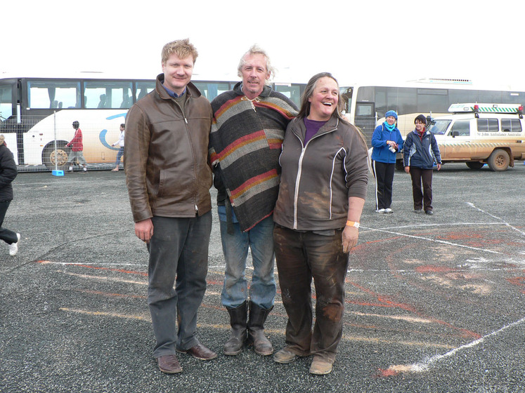 Three people stand together, there arms around each other, smiling at the camera. Around them on the bitumen ground are sand patterns and people observing the artwork.