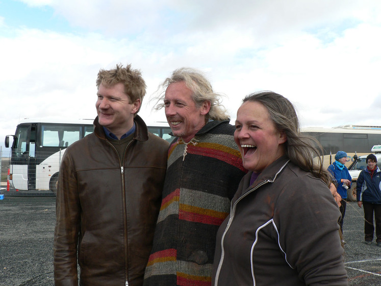 Three people stand together, there arms around each other, smiling at the camera. Around them on the bitumen ground are sand patterns and people observing the artwork.