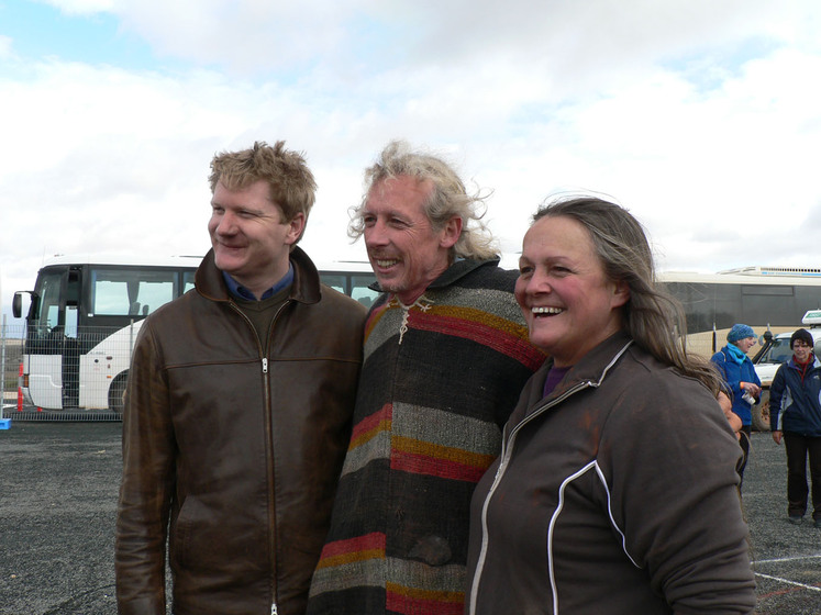 Three people stand together, there arms around each other, smiling at the camera. Around them on the bitumen ground are sand patterns and people observing the artwork.
