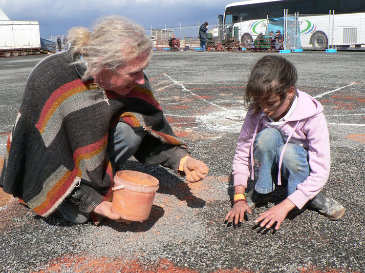 A person kneels on the ground, a fistful of sand in one hand and a bucket of red sand in the other. He is demonstrating to a young girl how to use the sand. The young girl has her hands hovered above the bitumen and is staring intensely at the ground.