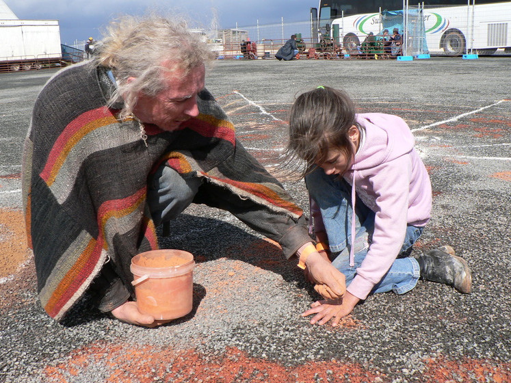 A person kneels on the ground, a fistful of sand in one hand and a bucket of red sand in the other. He is demonstrating to a young girl how to use the sand. The young girl has her hands pressed to the bitumen and is staring intensely at the ground.