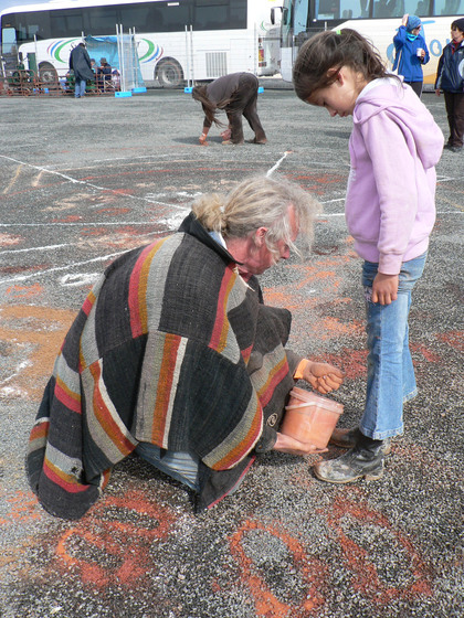 A person kneels on the ground, a fistful of sand in one hand and a bucket of red sand in the other. He is demonstrating to a young girl how to use the sand. The young girl is standing next to him, watching the person work.