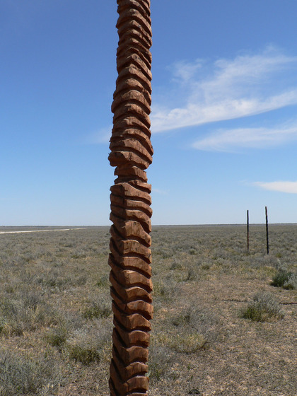 A wooden pole like sculpture, with small indents into the wood, almost making it look like stones stacked on one another. The landscape it is positioned in is dry, scrubby and sandy.