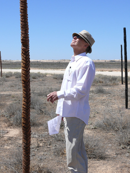 A man in a white lined shirt and hat stares up at the wooden pole sculpture.