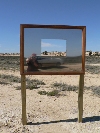 A large rectangle mirror stands on a wooden frame in the middle of a sandy scrubby landscape. It has a square cut out of the middle so you can see through to the view. In the mirrors reflection you can see someone taking a photograph, their car positioned behind them.