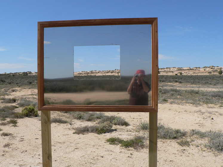 A large rectangle mirror stands on a wooden frame in the middle of a sandy scrubby landscape. It has a square cut out of the middle so you can see through to the view. In the mirrors reflection you can see someone taking a photograph, their car positioned behind them.