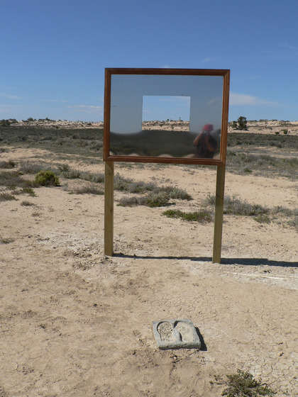 A large rectangle mirror stands on a wooden frame in the middle of a sandy scrubby landscape. It has a square cut out of the middle so you can see through to the view. In the mirrors reflection you can see someone taking a photograph, their car positioned behind them.