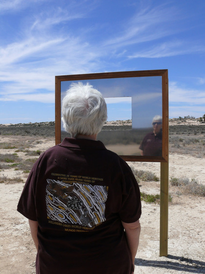 A woman, who's back is to the camera, faces a rectangle mirror positioned on a frame. The mirror is set in a sandy, scrubby landscape and has a small rectangle cut out of it so you can see through to the view. You can see the woman's reflection in the mirror.